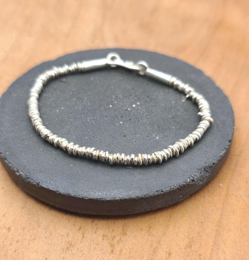 May include: A silver-toned bracelet composed of numerous small, thin, disc-shaped beads strung together in a continuous circular pattern. The bracelet features a simple metallic clasp mechanism at the top, connecting the two ends of the beaded strand.