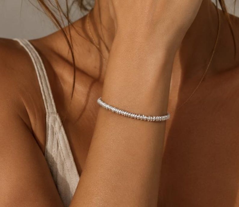 May include: A silver bracelet composed of numerous small, uniform, disc-shaped beads strung together in a continuous circular band. The metallic beads feature a reflective surface and are arranged tightly to create a textured, beaded appearance.