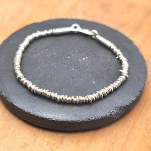 May include: A silver bracelet composed of numerous small, textured, disc-shaped beads strung together in a continuous circular pattern, featuring a metallic clasp mechanism at the top for securing the ends of the jewelry piece.