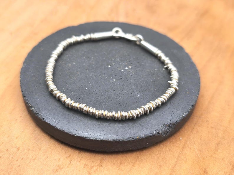May include: A silver bracelet composed of numerous small, textured, disc-shaped beads strung together in a continuous circular pattern, featuring a metallic clasp mechanism at the top for securing the ends of the jewelry piece.