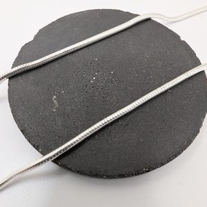 Op de afbeelding: Een zilverkleurige slangenketting met een gladde, flexibele en afgeronde schakelconstructie. De metalen ketting is gedrapeerd over een ronde, donkergrijze, gestructureerde, steenachtige displaybasis met een poreus oppervlak.