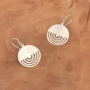 May include: A pair of silver-toned earrings. Each earring features a circular design with a half-circle cut-out pattern. The earrings have a brushed metal finish and are suspended from a simple hook.