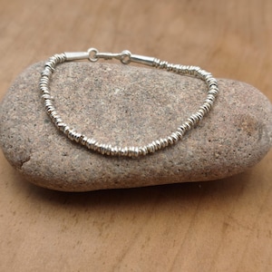 May include: A silver-colored bracelet composed of numerous small, thin, circular metallic discs strung together. The bracelet features a simple hook-and-eye clasp mechanism at the top, connecting the two ends of the continuous strand of beads.