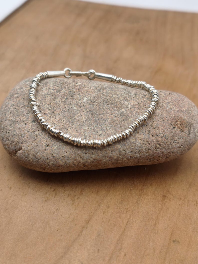 May include: A silver-colored bracelet composed of numerous small, thin, circular metallic discs strung together. The bracelet features a simple hook-and-eye clasp mechanism at the top, connecting the two ends of the continuous strand of beads.