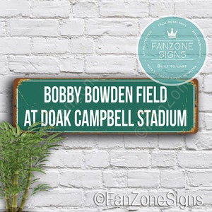 May include: A green and white metal sign with the text "BOBBY BOWDEN FIELD AT DOAK CAMPBELL STADIUM". The sign has a distressed look and is surrounded by a white brick wall.