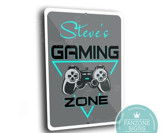 May include: A gray metal sign with a white border. The sign has a black and white illustration of a video game controller inside a triangle with the text "Steve's Gaming Zone" in black and turquoise blue lettering. The sign has a white crown logo with the text "FanZone Signs Built to Last" in black lettering.