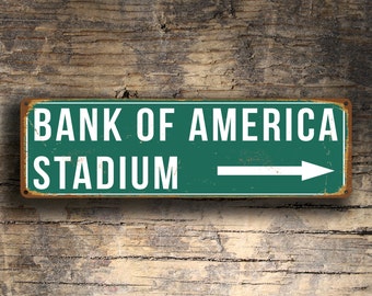 Op de afbeelding: Een groen metalen bord met witte tekst en een witte pijl die naar rechts wijst. Het bord staat er "Bank of America Stadium" op.