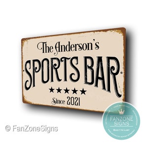 May include: A vintage-style metal sign with a distressed, rusty look. It reads "The Anderson's SPORTS BAR Since 2021" in black lettering on a cream background. The sign has five stars below the text.
