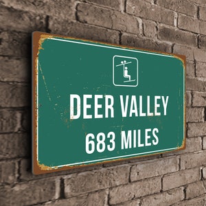 Könnte beinhalten: Ein grünes und braunes Vintage-Schild aus Metall mit weißem Text, der "DEER VALLEY 683 MILES" lautet. Oben links auf dem Schild befindet sich ein kleines weißes Symbol eines Skilifts.