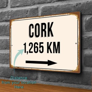 Könnte beinhalten: Rechteckiges Metallschild mit dem Wort "CORK" in fetten schwarzen Buchstaben und der Zahl "1.265 KM" darunter. Ein Pfeil zeigt nach rechts. Das Schild hat einen Used-Look.