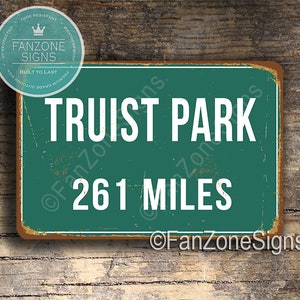 May include: A green metal sign with white text that reads "Truist Park 261 Miles". The sign has a distressed, vintage look and is surrounded by a brown border.