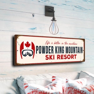 May include: A vintage-style metal sign with a red and white Canadian flag, a skier wearing goggles, and the text "Life is better in the mountains. Powder King Mountain Ski Resort."
