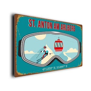 May include: A vintage-style metal sign with a teal background and the text "ST. ANTON AM ARLBERG" in red. The design features a skier, a gondola, and the coordinates 47.1333° N; 10.2667° E, all framed within a goggle shape.