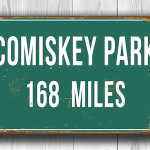 May include: A vintage-style metal sign with a green background and white text. The sign reads "Comiskey Park 168 Miles".
