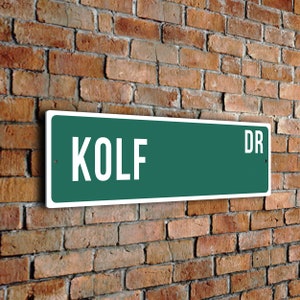 Può includere: Un cartello stradale verde con lettere bianche che recita "KOLF DR". Il cartello è montato su un muro di mattoni.