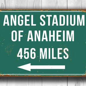 May include: A green and brown vintage-style metal sign with white text that reads "Angel Stadium of Anaheim 456 Miles" with a white arrow pointing to the left.