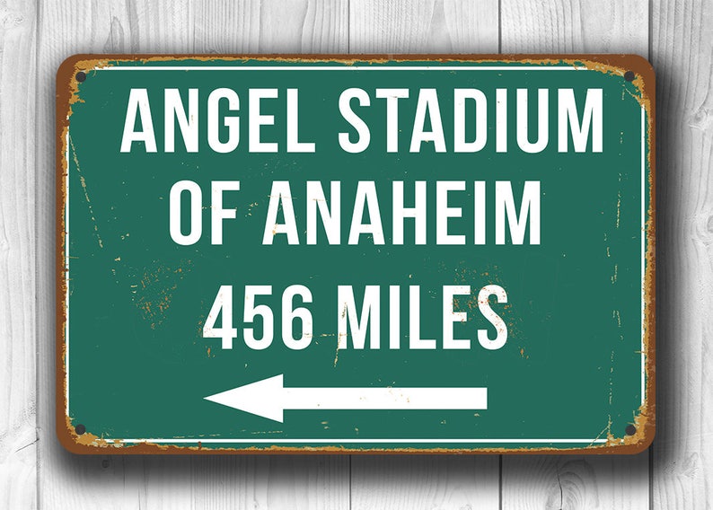 May include: A green and brown vintage-style metal sign with white text that reads "Angel Stadium of Anaheim 456 Miles" with a white arrow pointing to the left.