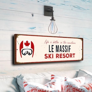 May include: A rectangular sign with a distressed, vintage appearance. It displays "LE MASSIF SKI RESORT" in red and navy blue, alongside a Canadian flag and ski goggles graphic. The phrase "life is better in the mountains" is also present.