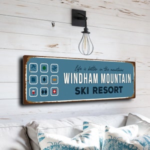 Könnte beinhalten: Ein blaues und braunes rechteckiges Schild mit weißem Text, der "Life is better in the mountains. Windham Mountain Ski Resort" lautet. Das Schild hat einen abgenutzten Look und zeigt verschiedene Ski-Symbole.
