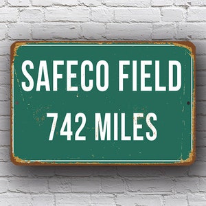 May include: A green metal sign with white text that reads "Safeco Field 742 Miles". The sign has a distressed, vintage look.