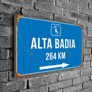 Op de afbeelding: Blauw en wit metalen bord met een skilift-icoon, de tekst "ALTA BADIA" en "264 KM" en een witte pijl die naar rechts wijst.