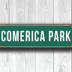 May include: A vintage-style metal sign with a green background and white lettering that reads "Comerica Park". The sign has a distressed, rusty look.