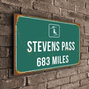 May include: A green and brown vintage-style metal sign with white text that reads "Stevens Pass 683 Miles". The sign features a small icon of a ski lift with two people riding it.