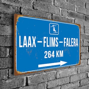 May include: A blue and white metal sign with a distressed look. The sign has a white arrow pointing to the right and the text "LAAX - FLIMS - FALERA 264 KM" in white. A small white icon of a chairlift is in the top right corner.