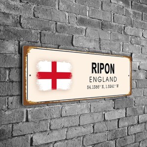 Pode incluir: Uma placa de metal retangular com um visual vintage e desgastado. A placa apresenta o texto "RIPON ENGLAND" em preto, juntamente com coordenadas. A bandeira inglesa está à esquerda. A placa está montada numa parede de tijolos cinzentos.