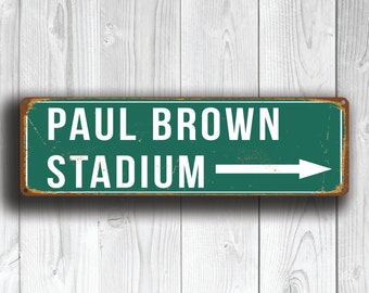 Pode incluir: Uma placa de metal verde e marrom com texto branco que diz "PAUL BROWN STADIUM" com uma seta branca apontando para a direita.