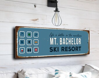 Könnte beinhalten: Ein blaues und braunes rechteckiges Schild mit dem Text "Life is better in the mountains. Mt. Bachelor Ski Resort." Das Schild hat einen abgenutzten Look und zeigt Symbole von Skifahrern, Snowboardern und Skiliften.