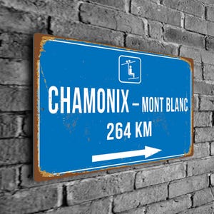 Op de afbeelding: Blauw en wit metalen bord met een skilift-icoon, de tekst "Chamonix - Mont Blanc" en "264 KM" en een witte pijl die naar rechts wijst.
