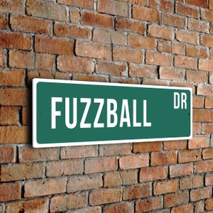 May include: A green street sign with white lettering that reads "Fuzzball Dr."