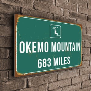 Könnte beinhalten: Ein grünes und braunes Metallschild mit einer weißen Silhouette eines Skilifts und dem Text "OKEMO MOUNTAIN 683 MILES".
