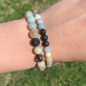 May include: Two bracelets stacked on a wrist. The top bracelet is made of light blue and green stones. The bottom bracelet is made of black lava rock and white stones.
