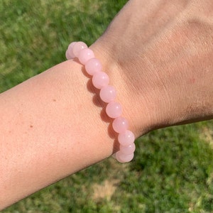May include: A pink rose quartz beaded bracelet on a person's wrist. The bracelet is made of round beads and has a simple design.