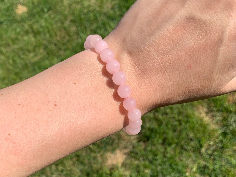 May include: A pink rose quartz beaded bracelet on a person's wrist. The bracelet is made of round beads and has a simple design.