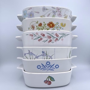 May include: A stack of six white ceramic casserole dishes with floral designs. Each dish has a different pattern, including flowers, tulips, and cherries. The dishes have handles on each side.