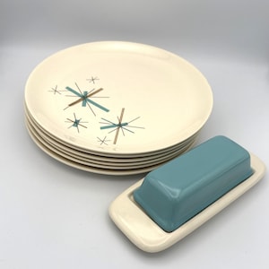 May include: A set of cream-colored ceramic plates with a mid-century modern design featuring teal and brown starburst patterns. A matching butter dish with a teal lid sits beside the plates. The dish has a cream-colored base.