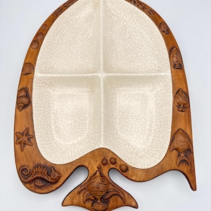 May include: A decorative serving dish with a leaf-like shape. The dish features four compartments with a cream-colored, cracked glaze. The outer rim is made of brown wood and is adorned with carved sea creatures, including starfish and seahorses.