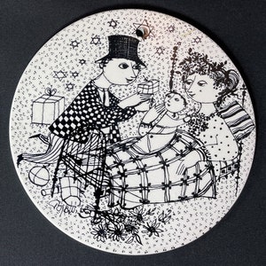 May include: Black and white illustration of a family with a baby receiving a gift. The illustration is on a round ceramic plate with a speckled background. The plate has a hole at the top for hanging.