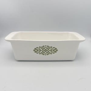 May include: A rectangular, off-white baking dish with a decorative olive green floral design on the front. The dish has a simple, classic design with slightly flared sides and handles. This vintage bakeware is ideal for baking and serving.