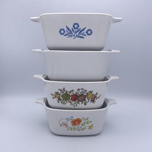May include: Four white rectangular casserole dishes stacked on top of each other. Each dish has a handle on either side. The top dish has a blue floral design. The second dish is plain. The third dish has a floral and fruit design. The bottom dish has a floral design.
