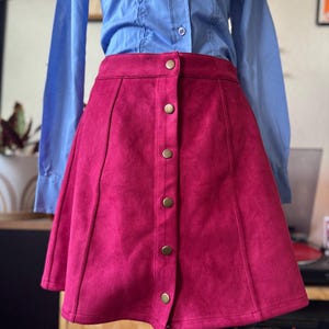 May include: A magenta-colored, button-down skirt with a flared A-line silhouette. The skirt features a series of gold-colored buttons down the front and is paired with a light blue button-down shirt. The skirt is made of a soft, suede-like material.