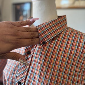 Puede incluir: Camisa a cuadros en naranja, turquesa, rojo y blanco, exhibida en un maniqu&iacute;. La camisa tiene un cuello cl&aacute;sico y botones negros. El dise&ntilde;o a cuadros es una caracter&iacute;stica destacada de la prenda.