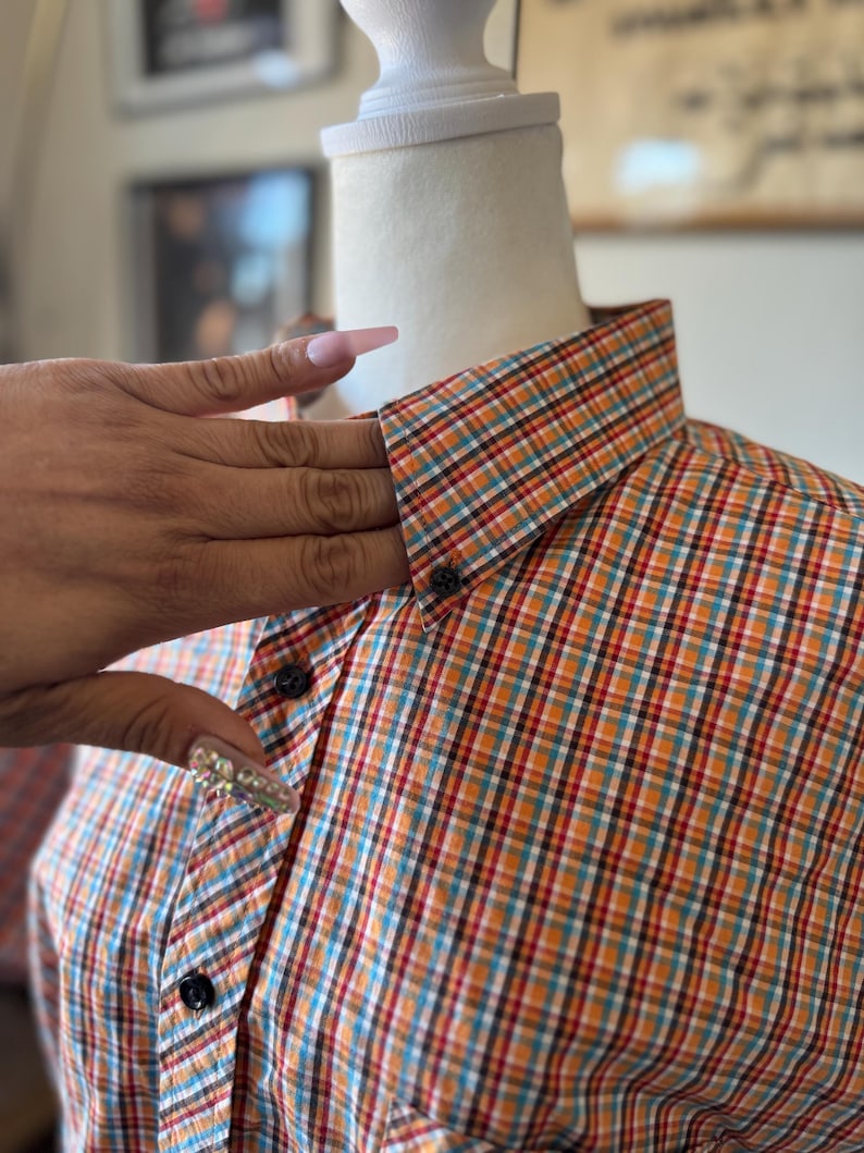 Puede incluir: Camisa a cuadros en naranja, turquesa, rojo y blanco, exhibida en un maniqu&iacute;. La camisa tiene un cuello cl&aacute;sico y botones negros. El dise&ntilde;o a cuadros es una caracter&iacute;stica destacada de la prenda.