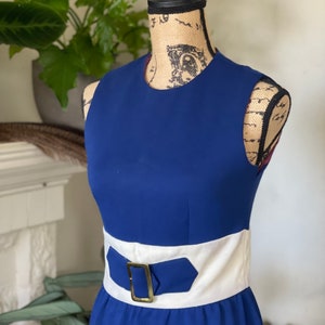 Peut inclure: Une robe bleue sans manches avec une ceinture blanche et une boucle dor&eacute;e. La robe a un design simple et classique.