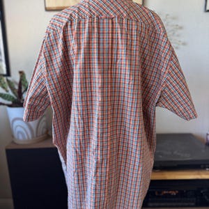 Puede incluir: Camisa de manga corta con botones en un patr&oacute;n de cuadros rojos, naranjas, azules y blancos. La camisa se muestra en un maniqu&iacute;, mostrando la parte posterior de la prenda. El cuello tiene un peque&ntilde;o bot&oacute;n oscuro.