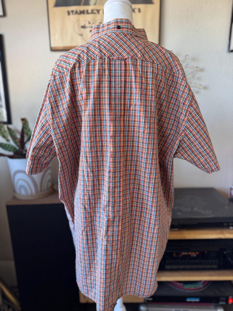 Puede incluir: Camisa de manga corta con botones en un patr&oacute;n de cuadros rojos, naranjas, azules y blancos. La camisa se muestra en un maniqu&iacute;, mostrando la parte posterior de la prenda. El cuello tiene un peque&ntilde;o bot&oacute;n oscuro.