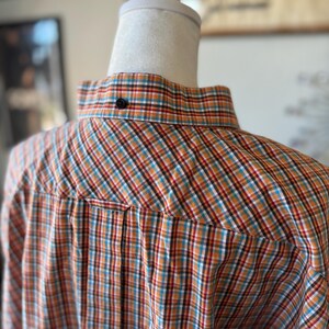 Puede incluir: Primer plano de una camisa abotonada con un estampado de cuadros en naranja, rojo, azul y blanco. La camisa se muestra en un maniqu&iacute;. El cuello tiene un solo bot&oacute;n negro.